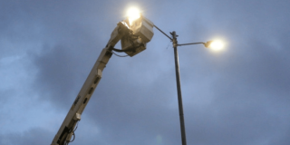 Poste de alumbrado público encendido mientras una plataforma elevadora realiza labores de mantenimiento en la parte superior, bajo un cielo nublado al atardecer.