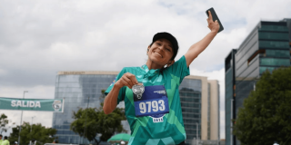 Participante de la Bimbo Global Race con camiseta verde y dorsal 9193 levanta un brazo mientras corre. Al fondo se observan edificaciones y el cielo despejado.
