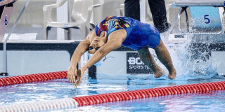 Nadador realiza la partida de espalda desde el borde de una piscina de competencia. La deportista viste traje de baño azul, gafas y gorro de natación, mientras se arquea hacia atrás para ingresar al agua.