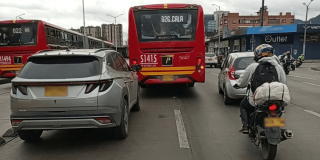 Bus articulado rojo de TransMilenio circula por una vía urbana junto a un automóvil gris y una motocicleta con carga en la parte trasera.