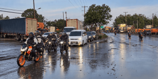 Motocicletas, vehículos particulares y camiones circulan por una vía mojada tras una lluvia, con reflejos sobre el pavimento y tránsito lento en ambos sentidos. Al fondo se observan árboles, postes de energía y señalización vial.