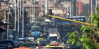 Panorámica de una vía con alta congestión vehicular en Bogotá. Se observan buses de TransMilenio, automóviles, motocicletas y vehículos de carga circulando en varios carriles, junto con agentes de tránsito en la calzada.