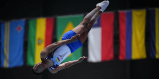 Deportista del Equipo Bogotá ejecuta un salto en competencia de atletismo, captado en el aire durante la prueba. Al fondo se observan banderas de distintos países participantes.