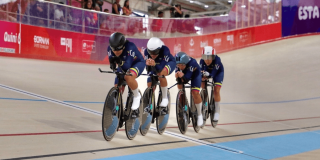 Cuatro ciclistas colombianas compiten en una pista de ciclismo de velódromo, avanzando en formación sobre sus bicicletas de pista mientras usan uniformes azul oscuro y cascos de protección.