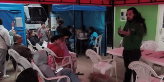 Personas sentadas en sillas plásticas asisten a una charla en un espacio cubierto, mientras una persona expone junto a una mesa.