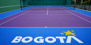Cancha de tenis con superficie morada y zonas laterales azules, delimitada por líneas blancas y una red en el centro. En el primer plano se observa el logo de Bogotá en color blanco y amarillo sobre la superficie de la cancha.