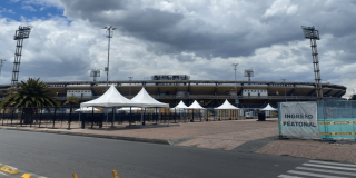 Vista exterior de una terminal de transporte con varias carpas blancas alineadas en la zona de acceso. Al fondo se observa un edificio de varios niveles y un cielo nublado con abundantes nubes grises. En primer plano hay una amplia superficie pavimentada sin presencia visible de personas.