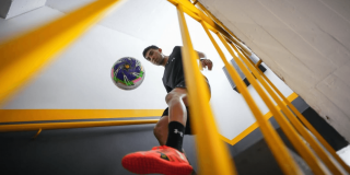 Messedinho realizando dominio de balón de fútbol en una escalera interior, vista desde abajo entre barandas amarillas que enmarcan la escena.