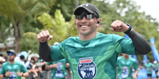 Participante de una carrera atlética viste camiseta verde de la Bimbo Global Race, gorra negra, gafas oscuras y audífonos inalámbricos, mientras avanza por el recorrido junto a otros corredores en un entorno al aire libre rodeado de árboles.