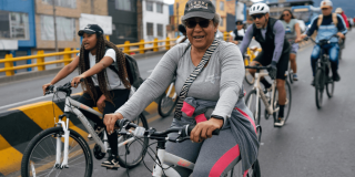 Personas en bicicleta recorren una vía urbana durante una jornada de Ciclovía. En primer plano, una persona vestida con ropa deportiva gris y gorra conduce una bicicleta blanca