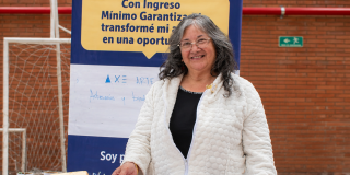 Ciudadana mayor posa en un stand de su emprendimiento en un evento organizado por la Secretaría Distrital de Integración Social, teniendo tras de ella un pendón sobre el programa Ingreso Mínimo Garantizado (IMG).