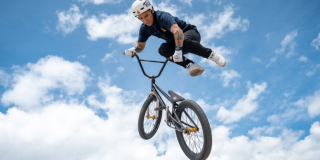Luis Oswaldo Rincón ejecuta una maniobra aérea en una bicicleta BMX durante una competencia de ciclismo extremo. La bicicleta se encuentra inclinada en el aire y el deportista viste casco, camiseta oscura y pantalón claro.