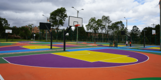 Cancha deportiva multicolor con tablero y aro de baloncesto, rodeada por cerramiento metálico, árboles y cielo nublado al fondo.