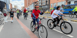 Ciclistas y peatones transitan por una vía urbana durante una jornada recreativa. En primer plano, dos personas montan bicicleta con casco mientras otras personas avanzan en bicicleta y caminando por la calzada.