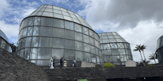 Vista exterior de los domos de vidrio del Tropicario del Jardín Botánico de Bogotá. En primer plano se observa un sendero elevado por el que transitan varias personas, mientras al fondo se aprecia un cielo parcialmente nublado y vegetación alrededor de la estructura.