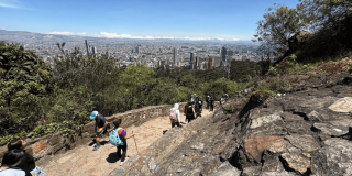 Personas recorren el sendero peatonal de Monserrate entre rocas y vegetación; al fondo se observa una vista panorámica de Bogotá bajo cielo despejado.