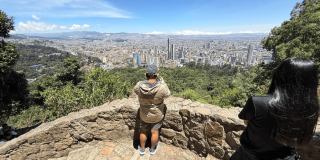 Vista panorámica de Bogotá desde un mirador en Monserrate. En primer plano se observa un muro de piedra y dos visitantes de espaldas contemplando la ciudad.