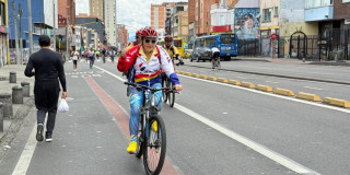 Persona con camiseta blanca, roja y azul monta una bicicleta sobre una vía de la Ciclovía de Bogotá; al fondo se observan edificaciones, vehículos y otros ciclistas.