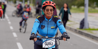 Participante de la Ciclovía de Bogotá se desplaza en bicicleta por una vía habilitada para la actividad. Lleva casco rojo, gafas y una camiseta deportiva azul con el número 0382 en la parte frontal.