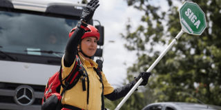 Persona con casco rojo, guantes y uniforme amarillo sostiene una paleta de señalización con la palabra “SIGA” mientras levanta una mano.