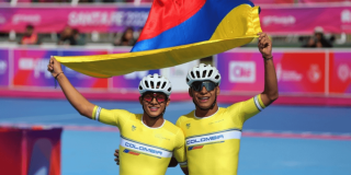 Dos deportistas de patinaje de velocidad con uniformes amarillos de Colombia y cascos blancos sostienen una bandera de Colombia sobre sus cabezas en una pista de competencia.