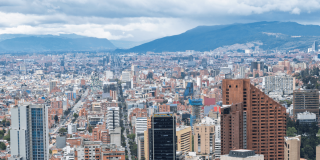 Vista panorámica de Bogotá con edificios residenciales y empresariales, avenidas principales y cerros al fondo bajo un cielo cubierto de nubes grises.