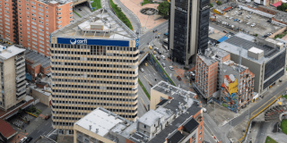 Vista aérea de edificios y vías en el centro de Bogotá. En primer plano destaca un edificio con el letrero “Corfi Colombiana”, rodeado de torres de oficinas, calles con vehículos en circulación y un mural de gran formato en la fachada de un edificio cercano.