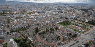 Vista panorámica de Bogotá con edificaciones residenciales y comerciales, corredor vial rodeado de árboles, amplias zonas verdes, cementerio y conjuntos de apartamentos al fondo bajo cielo nublado.