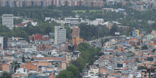 Vista panorámica de Bogotá con edificaciones residenciales y comerciales, corredor vial rodeado de árboles, amplias zonas verdes y conjuntos de apartamentos al fondo bajo cielo nublado.