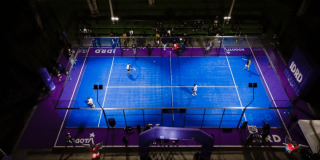 Vista aérea de varias canchas de pádel iluminadas en color azul dentro de un complejo deportivo cubierto, con jugadores en acción y áreas de circulación alrededor de los escenarios deportivos.