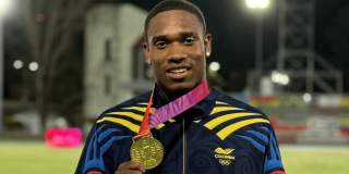 Ronal Longa sostiene una medalla dorada frente a una pista de atletismo iluminada durante la noche. En la prenda se observa el logo del Comité Olímpico Colombiano.