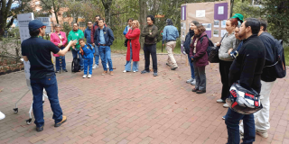 Personas participan en una actividad al aire libre sobre una plazoleta de ladrillo. Varias están alineadas y observan una demostración; algunas llevan chaquetas azules y una persona en el centro viste una chaqueta roja.