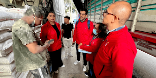 Personal con chaquetas rojas realiza una visita de inspección en una bodega de alimentos. Varias personas conversan entre pilas de sacos almacenados y un camión de carga estacionado junto al área de trabajo.