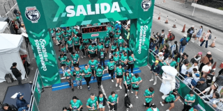 Cientos de participantes cruzan el arco de salida de la Bimbo Global Race 2026 sobre una vía urbana. El pórtico verde muestra el texto “SALIDA”, los logotipos de Bimbo Global Race 2025 y la frase “COMPARTIMOS LO BUENO”. A los lados hay espectadores detrás de vallas, mientras corredores con camisetas verdes avanzan por la línea de partida.
