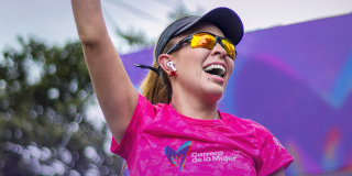 Participante de la Carrera de la Mujer con camiseta rosada, gorra negra, gafas deportivas y audífonos inalámbricos, levantando un brazo durante una actividad del evento al aire libre.