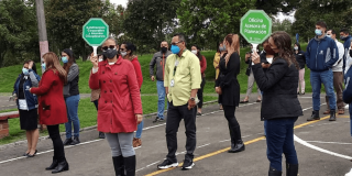 Grupo de personas participando en una actividad de evacuación al aire libre, posiblemente un simulacro de emergencia. Algunas llevan tapabocas y sostienen señales con los nombres de diferentes dependencias institucionales para el Simulacro Bogotá 2026.