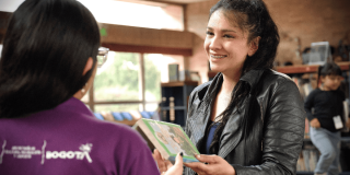 Imagen de una mujer sonriendo mientras entrega un libro a funcionaria de BibloRed de espaldas.