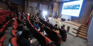 Asistentes participan en una jornada de socialización del Plan de Acción Acústico de Bogotá en un auditorio, mientras una presentación se proyecta en pantalla principal.