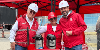 Alcalde Mayor de Bogotá, Carlos Fernando Galán, junto a Leonidas Narváez, gerente de la Línea 1 del Metro de Bogotá y Neida Ospina, vendedora de café en el carro ambulante 'Metro Tinto' visten con chalecos y cascos de seguridad rojos y blancos permanecen bajo una carpa roja junto a una mesa con termos de café. En las prendas se observan los logos de Bogotá y Metro de Bogotá.