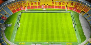 Vista general de la cancha del estadio El Campín con césped verde cortado en franjas longitudinales. En la parte superior se observan graderías con sillas amarillas y rojas, una franja publicitaria del IDRD y parte de la cubierta del escenario deportivo.