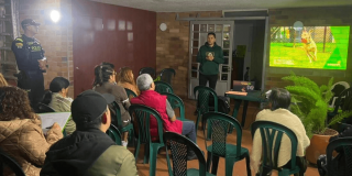 Charla informativa sobre el cuidado de animales de compañía en un salón comunitario. Varias personas sentadas en sillas de plástico observan una presentación proyectada en pantalla, mientras una persona expone al frente y un agente de policía permanece de pie al lateral con proyección en pared de un perro en zona verde.