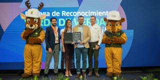 Entrega de reconocimiento ambiental a una entidad distrital. En una tarima, varias personas sostienen un galardón mientras posan para una fotografía, acompañadas por una mascota institucional. Al fondo se observa una pantalla azul con texto relacionado con el programa "Sello Verde de Verdad".