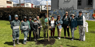 Grupo de personas reunidas alrededor de un árbol recién sembrado en una zona verde de un parque. Varias personas sostienen herramientas de jardinería mientras, al fondo, se observan edificaciones residenciales y montañas bajo un cielo parcialmente nublado.