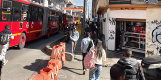 Peatones caminando por una acera junto a autobuses articulados rojos de TransMilenio en una concurrida vía urbana de Bogotá.