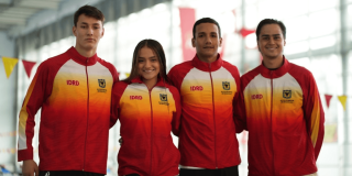 Cuatro deportistas del Equipo Bogotá posan juntos en una instalación deportiva cubierta. Llevan chaquetas de equipo con degradado rojo, naranja y amarillo, cremallera frontal y los textos visibles ‘IDRD’ y ‘Alcaldía Mayor de Bogotá D.C.’ en el pecho.