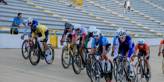 Grupo de ciclistas compite en una pista inclinada de velódromo durante una prueba de ciclismo. Las y los deportistas avanzan en fila sobre bicicletas de ruta, con uniformes de distintos colores y cascos de competición, mientras recorren la curva de la pista de madera.