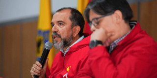 Primer plano de una mesa de reunión institucional. El director del IDRD, Daniel García Cañón, con chaqueta roja sostiene un micrófono y habla, mientras otra persona, también con chaqueta roja, aparece desenfocada en primer plano durante rueda de prensa para explicar situación de la gramilla del estadio El Campín de Bogotá.