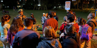 Grupo de personas reunidas en un parque durante una jornada nocturna de preparación para simulacro. Un integrante del cuerpo de bomberos brinda indicaciones mientras una persona sostiene un letrero con la identificación "Edificio B5".