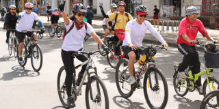 Personas participan en la Ciclovía de Bogotá recorriendo una vía urbana en bicicleta. En primer plano, varias personas usan casco y pedalean por la calzada, mientras otras ciclistas avanzan detrás.