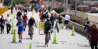 Personas caminan, trotan y montan bicicleta por una vía de la Ciclovía de Bogotá, delimitada con conos, mientras otras permanecen a los costados del recorrido.
